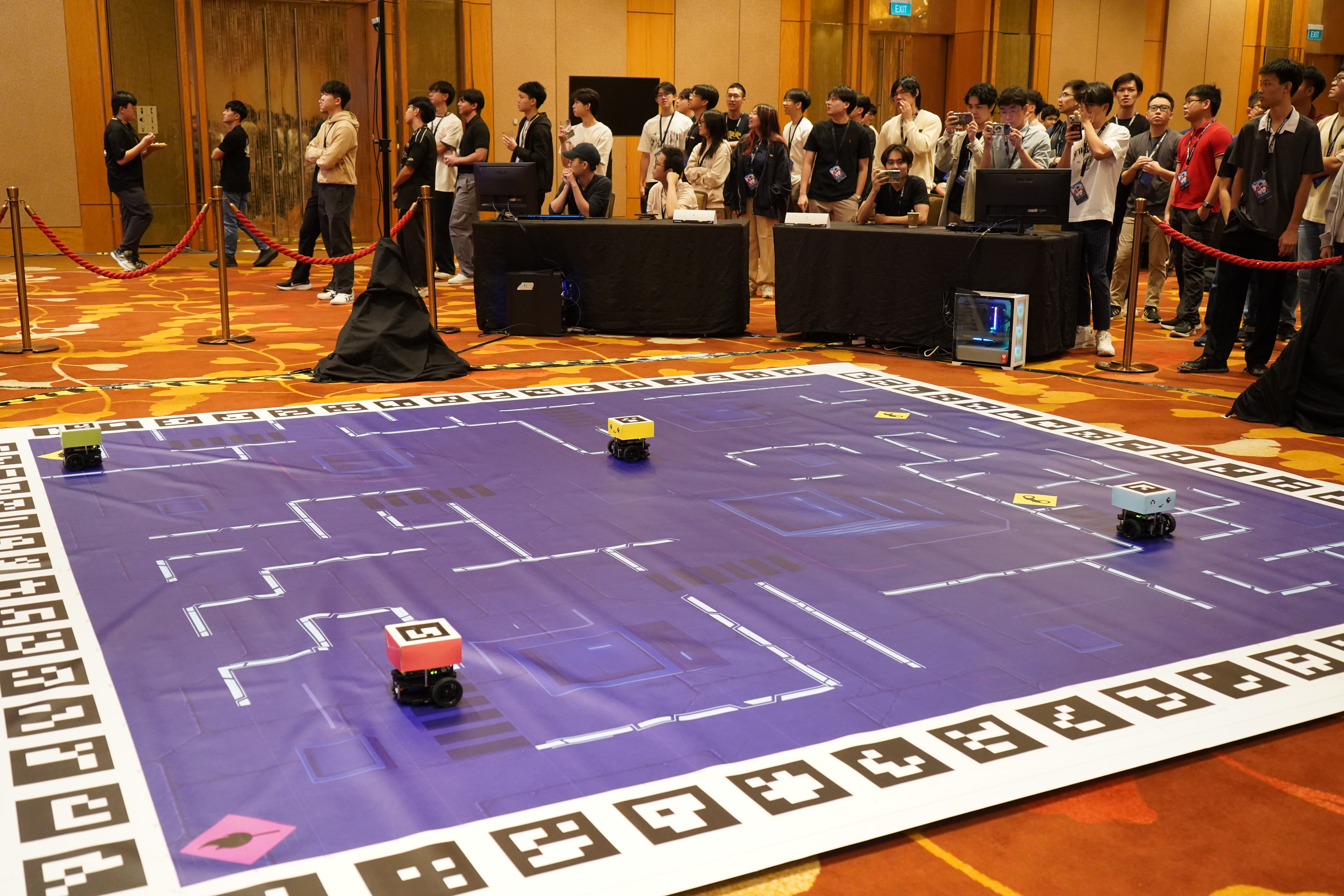 Snapshot captured of the robots moving around during the in-personal Finals. Four robots are in various locations around the arena, while a crowd of students watches on.